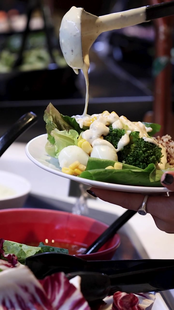 Un mestolo versa condimento bianco cremoso su un piatto di verdure fresche.