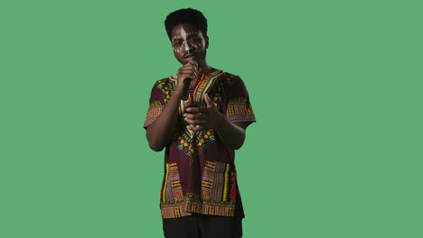 retrato de un joven afroamericano cantando una canción en el micrófono. hombre negro con un patrón étnico blanco en la cara y con una camisa brillante con ornamento nacional posando en la pantalla verde en el estudio