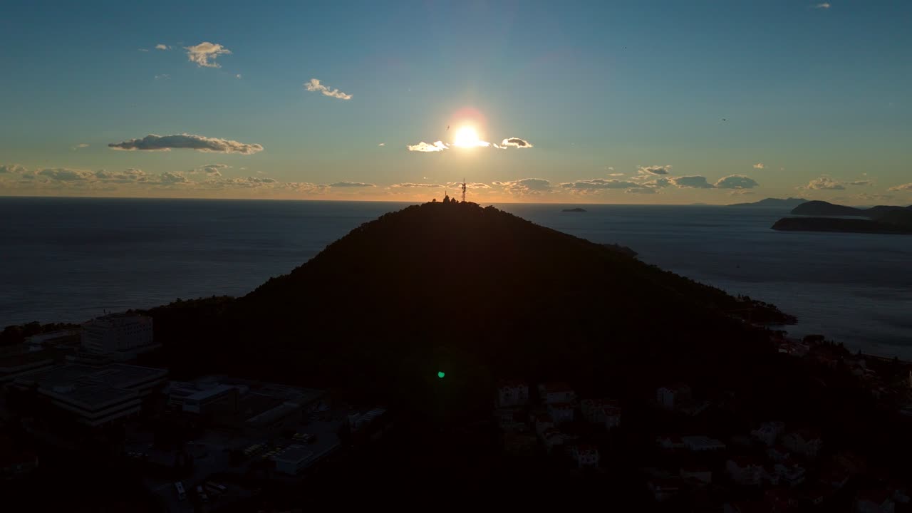 Aerial drone shot staring into the Mediterranean sunset at golden hour, with a glowing mountain silhouette in frame, soft orange sky reflecting over the calm blue sea and tranquil coastal landscape