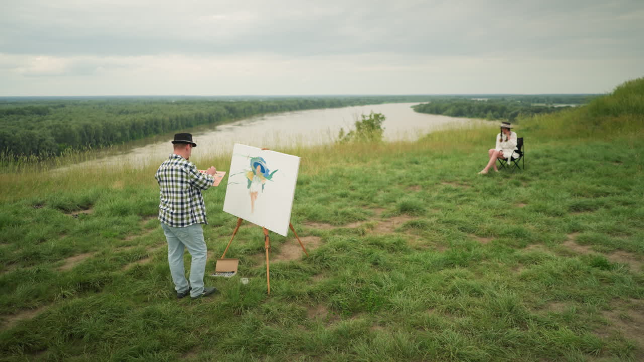 A skilled man, in a hat, checkered shirt, and jeans, is focused on creating a masterpiece on a canvas in a lush grassy field beside a lake. a woman in a hat and white dress sits comfortably on a chair