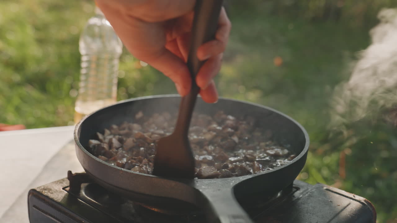 En campingstøbejernspande syder over en bærbar brænder. Et nærbillede viser en træspatel, der rører kødterninger i olie og krydderier, mens damp og røg stiger op mod en grøn campingpladsbaggrund, og en rig aroma spreder sig.
