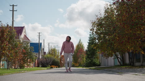 business woman in pink jacket and gray sweatpants walks away with paper bag swaying down residential street, surrounded by vibrant autumn foliage and quaint houses under bright skies