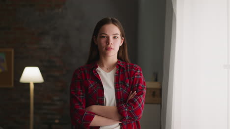 teenage girl with crossed arms stands near window at home