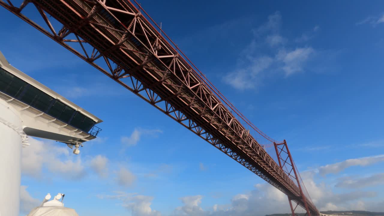 Dramatic low-angle view sailing under the iconic 25 de Abril suspension bridge in Lisbon, Portugal. Red steel truss structure against vivid blue sky from cruise ship deck
