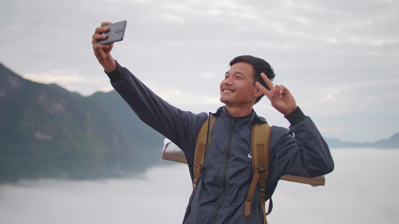 Asian Hiker Male Doing Victory Sign And Using Smartphone Taking Selfie Photo With The View Of Top Foggy Mountain