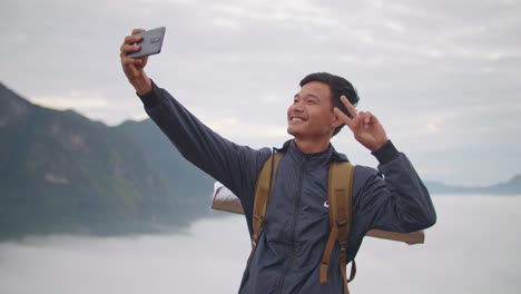 asian hiker male doing victory sign and using smartphone taking selfie photo with the view of top foggy mountain