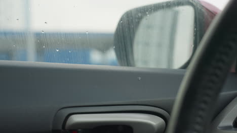 car window slides upward as snowflakes swirl outside against blurred building background visible through glass surface interior features steering wheel and door handle in foreground