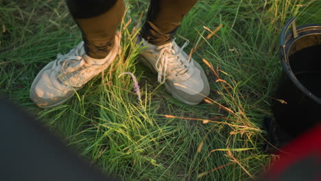 a close-up of legs in black trousers and white sneakers, dancing lightly in the grass near a black bucket