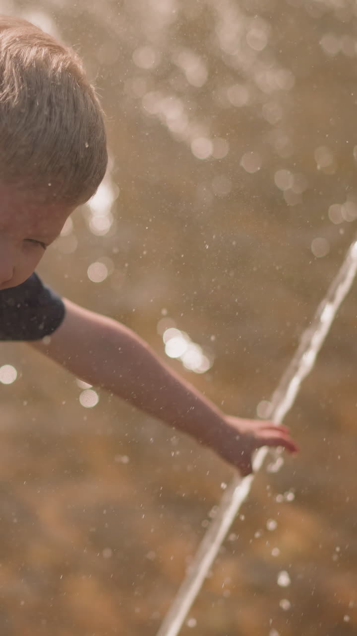 Joyful little child touches fountain water jet holding mother hand on square slow motion. Boy with parent has fun on summer day in hot weather