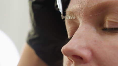 close-up of client with closed eyes during eyebrow treatment as technician gently applies mixed product to brow using angled brush, technician hand and ring light visible in blurred background