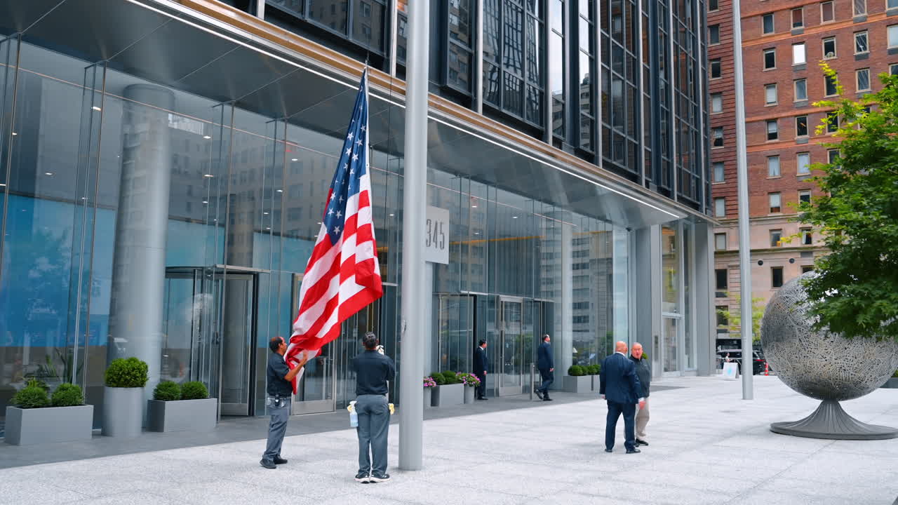 New York, USA, 2 ottobre 2025: Due uomini stanno abbassando la bandiera dall'asta all'aperto. Una grande installazione a sfera metallica si trova di fronte all'edificio. New York, USA