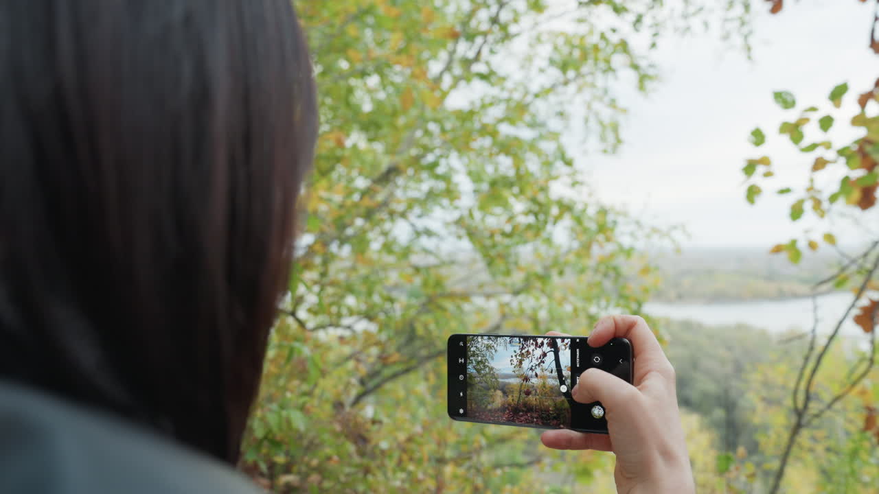 encuadre con teléfono sobre el hombro vista del río senderista con chaqueta de cuero sujeta un smartphone a la altura de los ojos, compone una toma panorámica del río y los abedules, pulsa el obturador, revisa la pantalla, ambiente otoñal melancólico