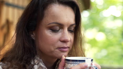 close up fhd shot of a middle-aged brunette woman enjoying the smell of a fresh cup of hot tea for breakfast in front of a weekend house in nature