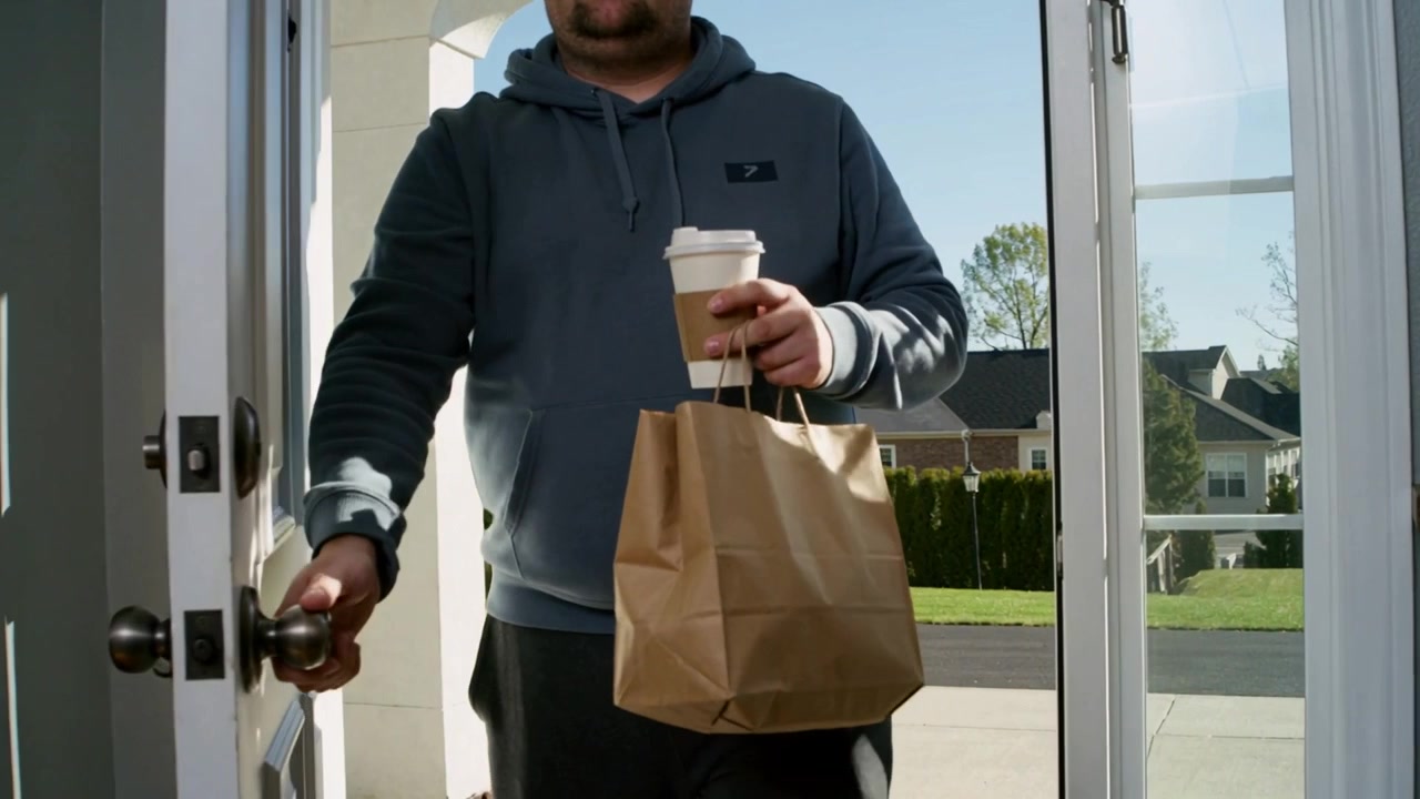 Approaching porch, adult male in gray hoodie turning doorknob bringing paper bag and coffee inside