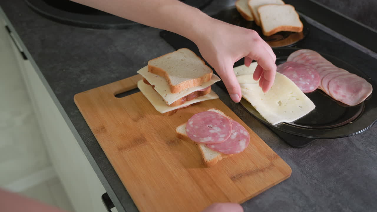 Hand placing slice of deli meat on sandwich stack with bread and cheese during meal preparation on wooden cutting board, highlighting homemade cooking, lunch routine