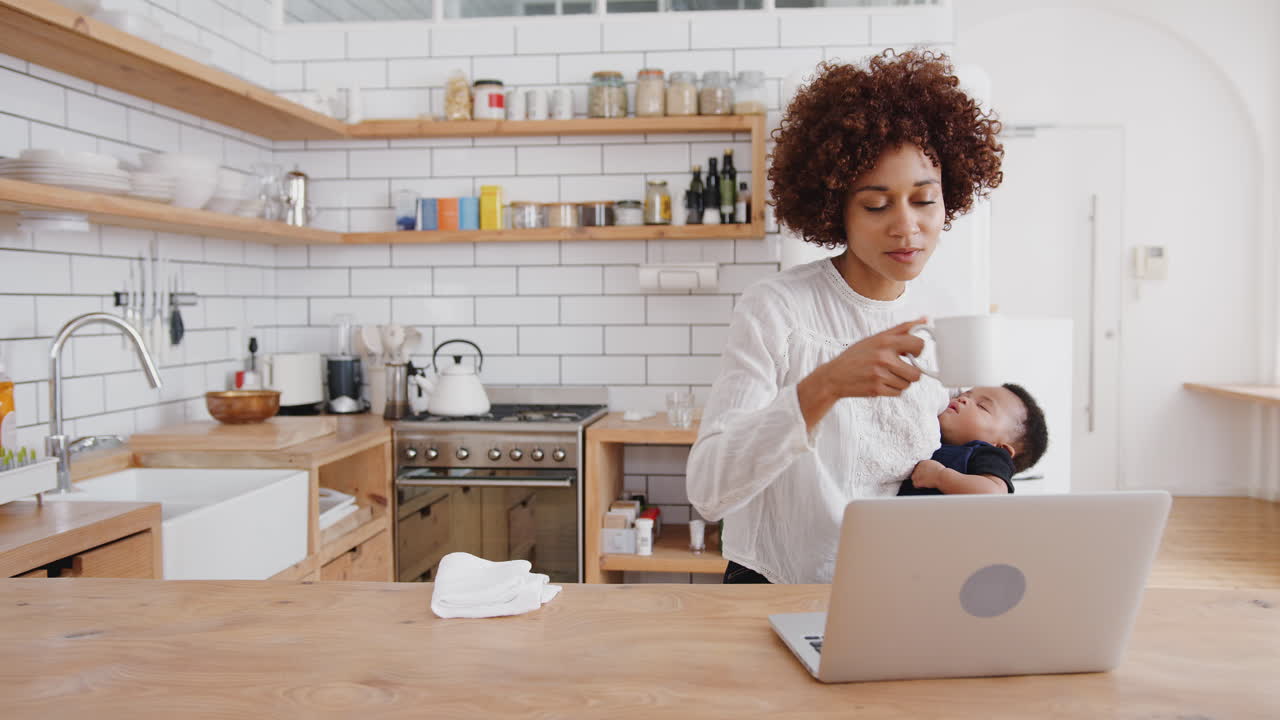 madre multitarea sostiene a su hijo bebé durmiendo mientras trabaja en la computadora portátil en la cocina