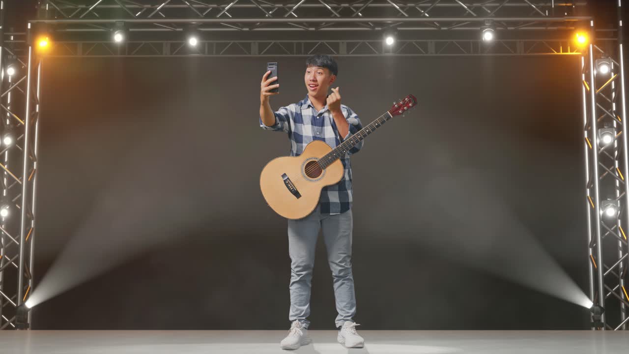 cuerpo lleno de adolescente asiático con selfie de guitarra en el teléfono inteligente mientras está de pie en el escenario