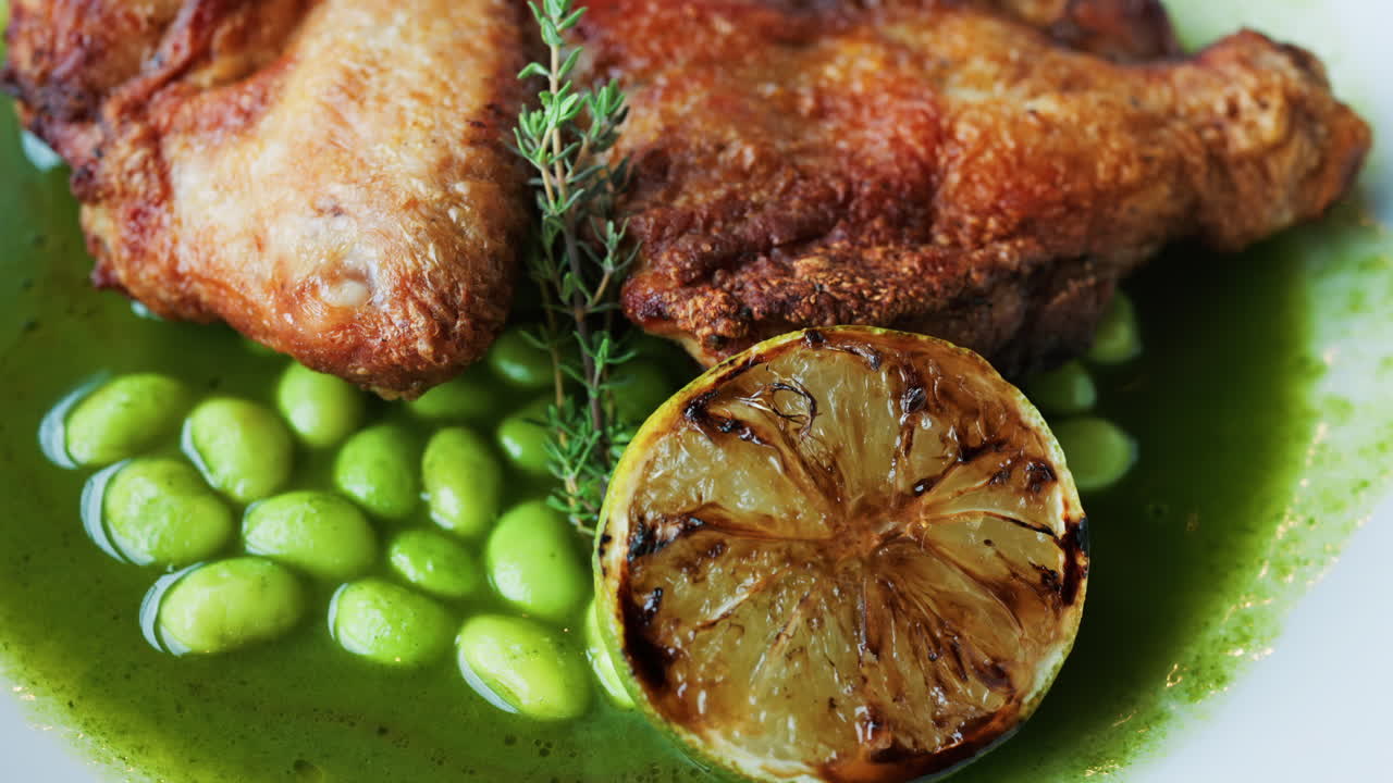 Close-up of a gourmet roasted chicken thigh served with edamame beans, a drizzle of herb-infused green sauce, and a charred lime half