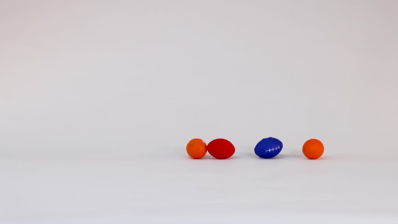 Small blue, orange, and red foam footballs bounce and roll across a white studio surface