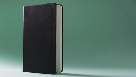 Close-up-of-closed-black-book-standing-vertical-with-copy-space-on-green-background-in-slow-motion