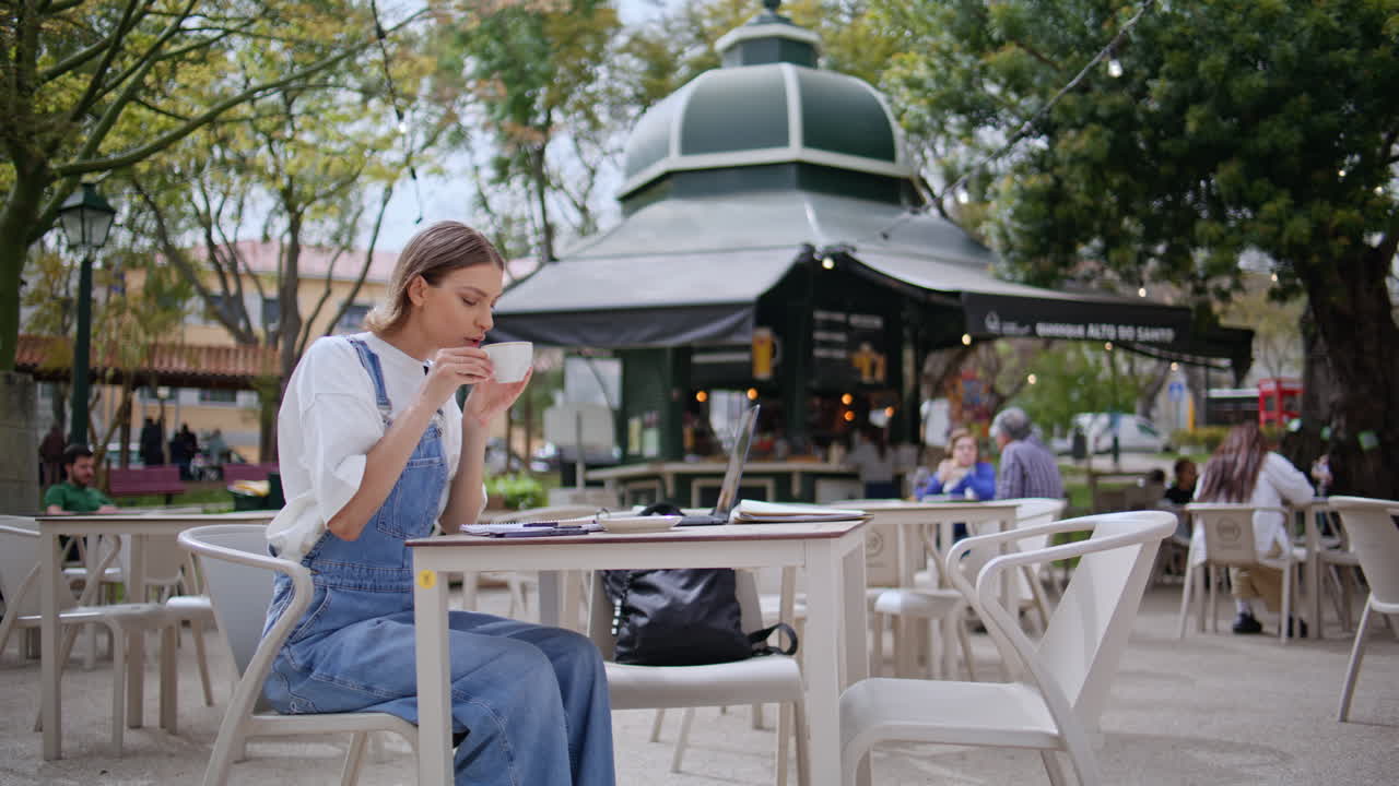 Smiling freelancer drinking cappuccino at cafeteria terrace. Woman typing laptop