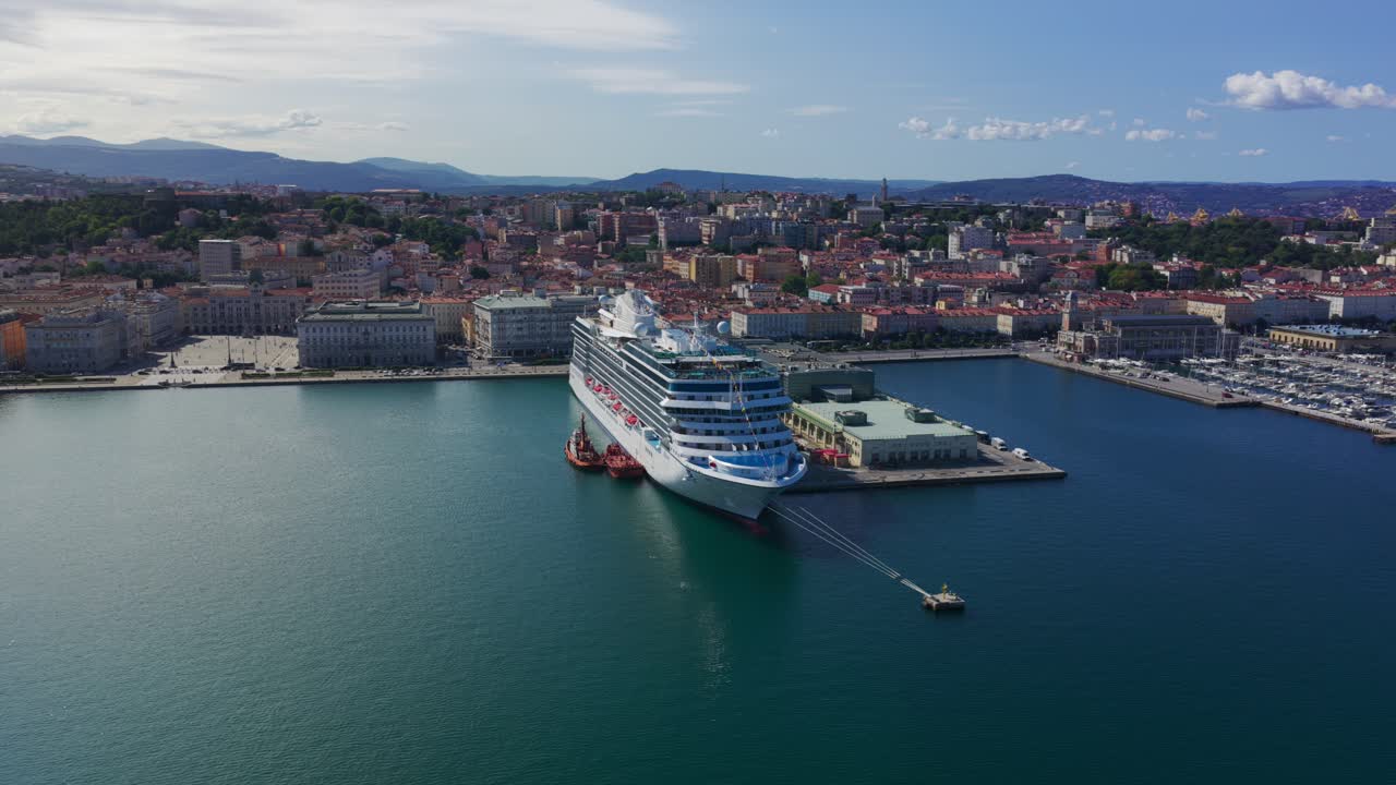 Aerial view of large cruise ship docked in port. Tugboats assist vessel. Cityscape of Trieste in background. For travel or transportation content, Italy