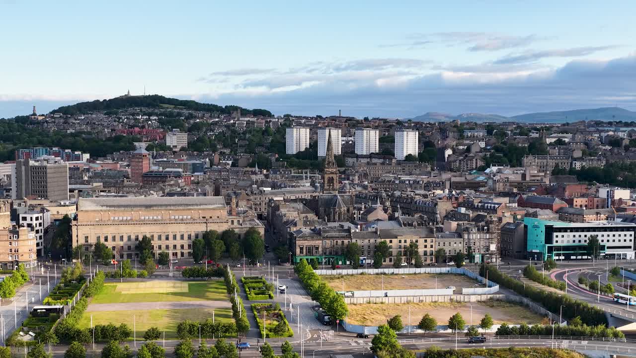 Aerial pan reveals Dundee’s riverfront, urban park, historic architecture, and distant hills in daylight