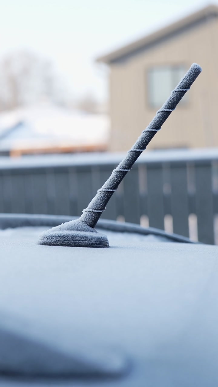 Frost-covered car antenna on a vehicle roof in a winter morning, shallow depth with suburban house blur. Cold weather texture detail for seasonal automotive b-roll.