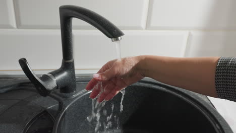 close up of woman in plaid patterned dress rinsing hand under flowing water from black kitchen faucet, with water bubbles visible on hand, focusing on hygiene