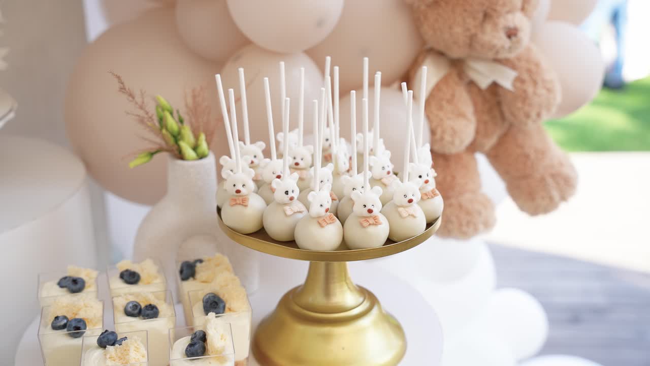 Elegante allestimento di tavolo da dessert con dolci bianchi e dorati, tra cui cake pop a forma di orsetto, mini cheesecake e una torta piumata, ideale per feste di compleanno o baby shower