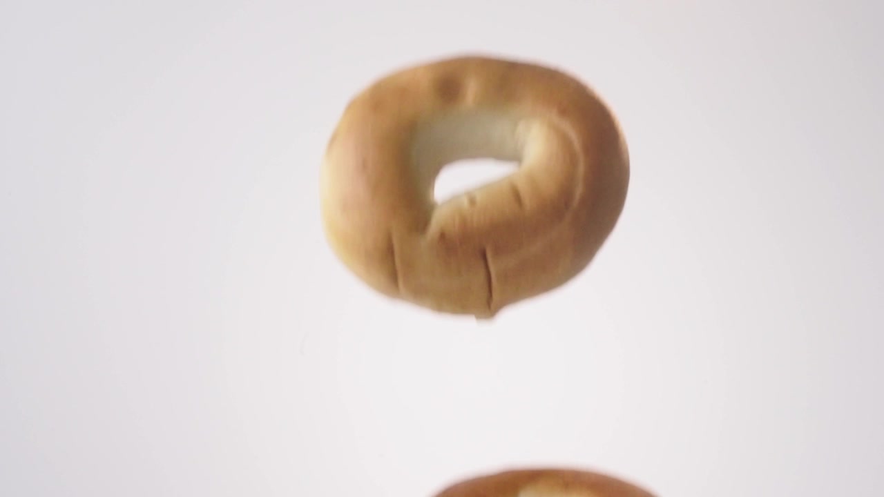 Chewy golden bagels cascading and falling through frame on white in slow motion. breakfast catering posts, hotel buffet content, coffee shop pastry videos.