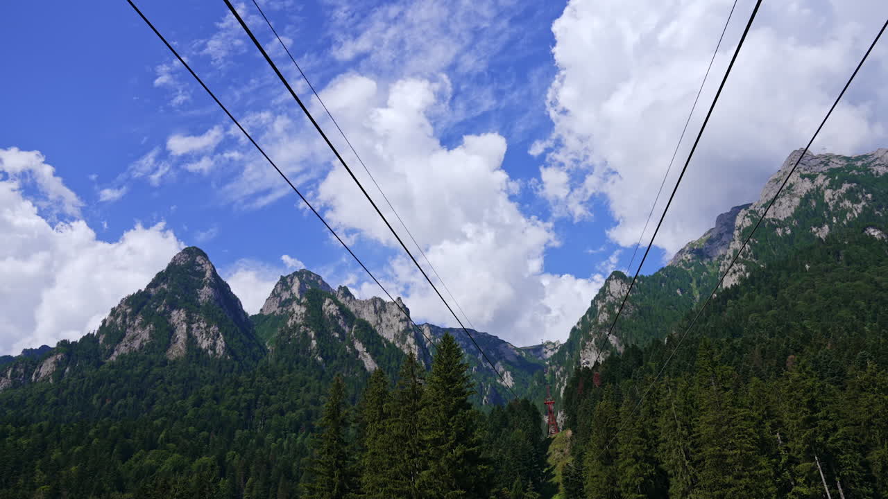 Majestic mountain range under blue sky. Clouds drift across a vibrant blue sky above towering mountains, surrounded by lush green trees and scenic beauty