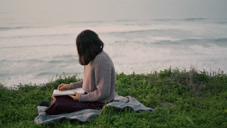 Mujer-Sentada-Junto-Al-Mar-Manta-Leyendo-Un-Libro-Con-Espectaculares-Vistas-Al-Mar.-Niña-Descansando.