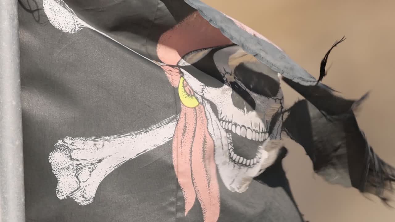 A weathered black pirate flag with skull and crossbones flutters against a blurred background