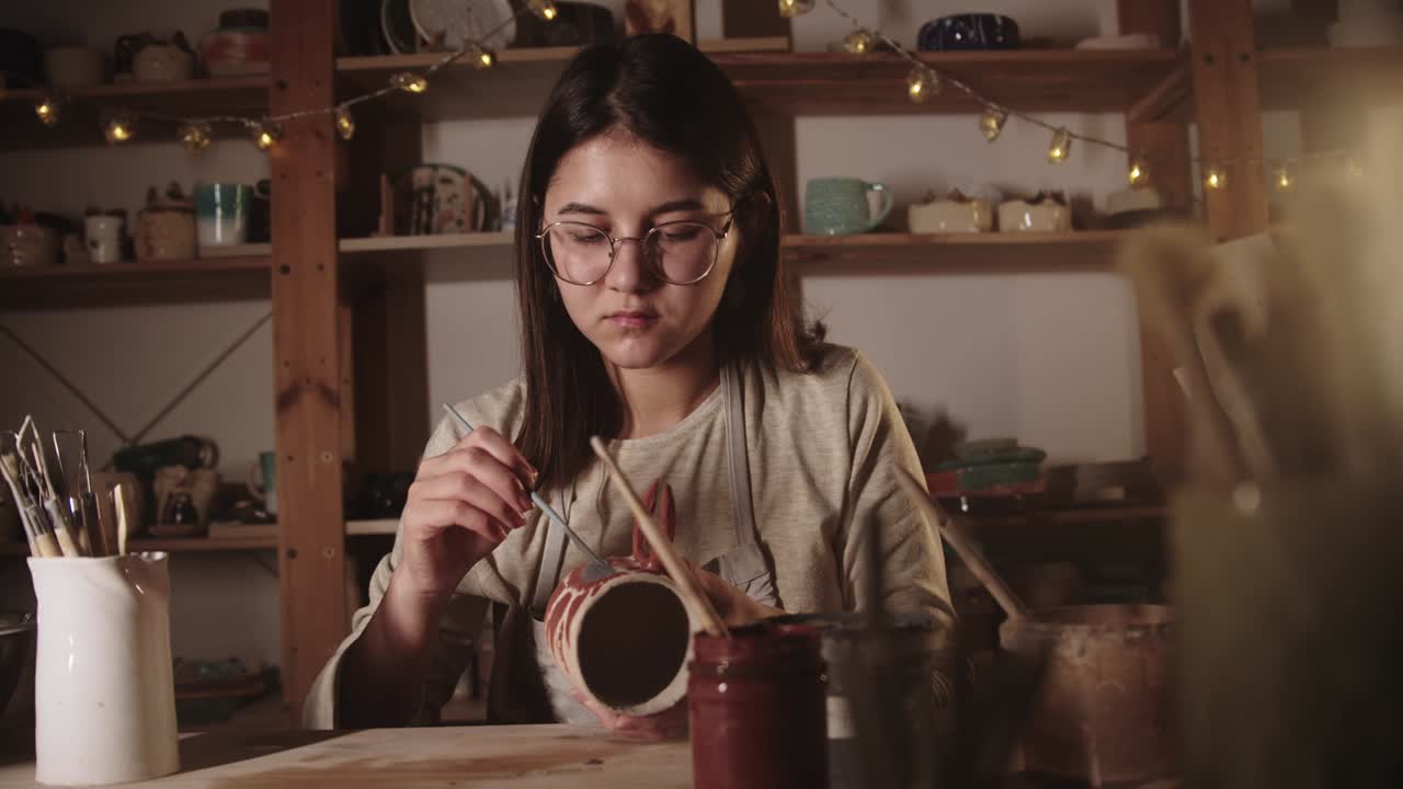 joven alfarera con gafas dibujando un diseño en la taza de cerámica con un pincel en un estudio de arte