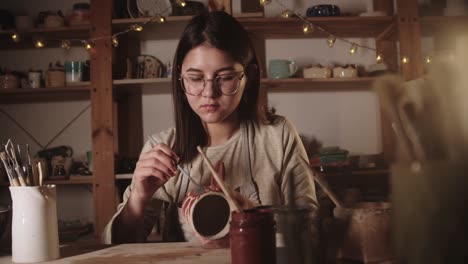 young woman potter in glasses drawing a design on the ceramic mug with a brush in art studio