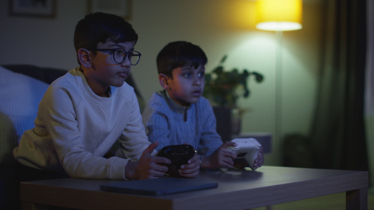 Two Young Boys At Home Playing With Computer Games Console On TV Holding Controllers Late At Night 7
