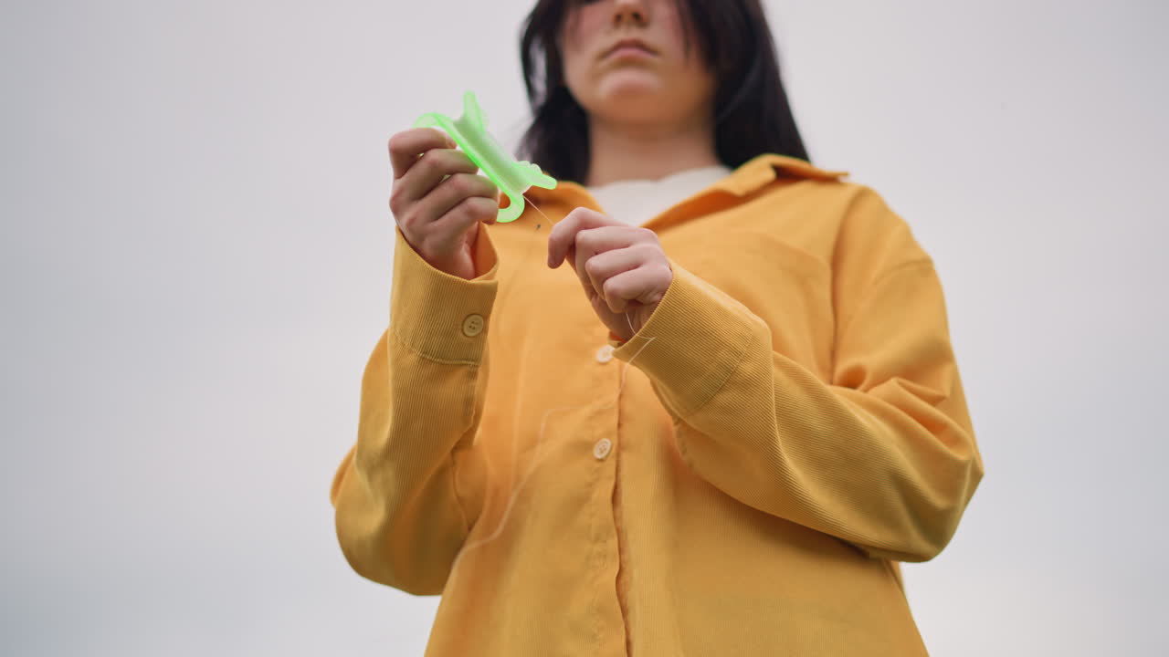 woman yellow jacket holding green toy, lowangle outdoor portrait under overcast sky, contemplative lone figure fidgets with plastic clip, textured fabric and button details, quiet cinematic mood