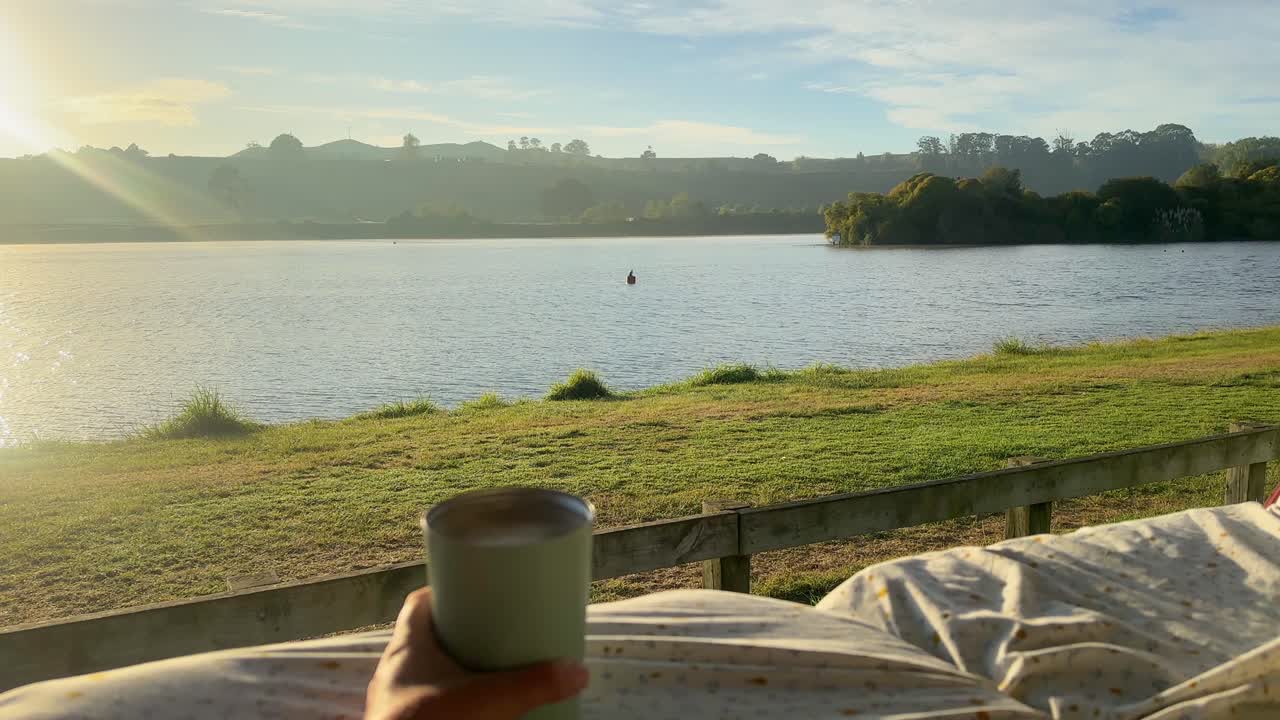 A POV shot of enjoying a coffee from the back of a camper van overlooking a stunning lake in New Zealand. Calm waters, lush greenery, and peaceful surroundings make for a tranquil moment