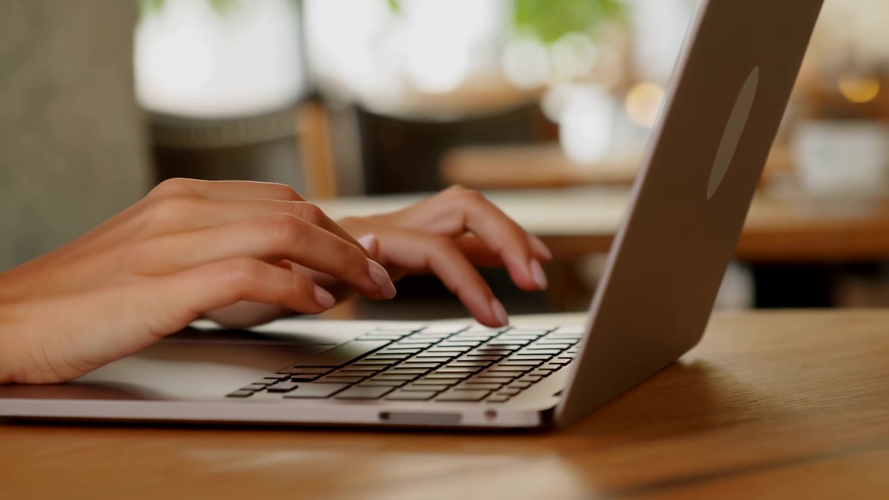 Dita in primo piano si muovono sui tasti del laptop mentre il dispositivo poggia su una superficie di legno liscia, con una luce brillante dalla finestra e sedie leggermente sfocate sullo sfondo che enfatizzano l'interazione costante con la tastiera.