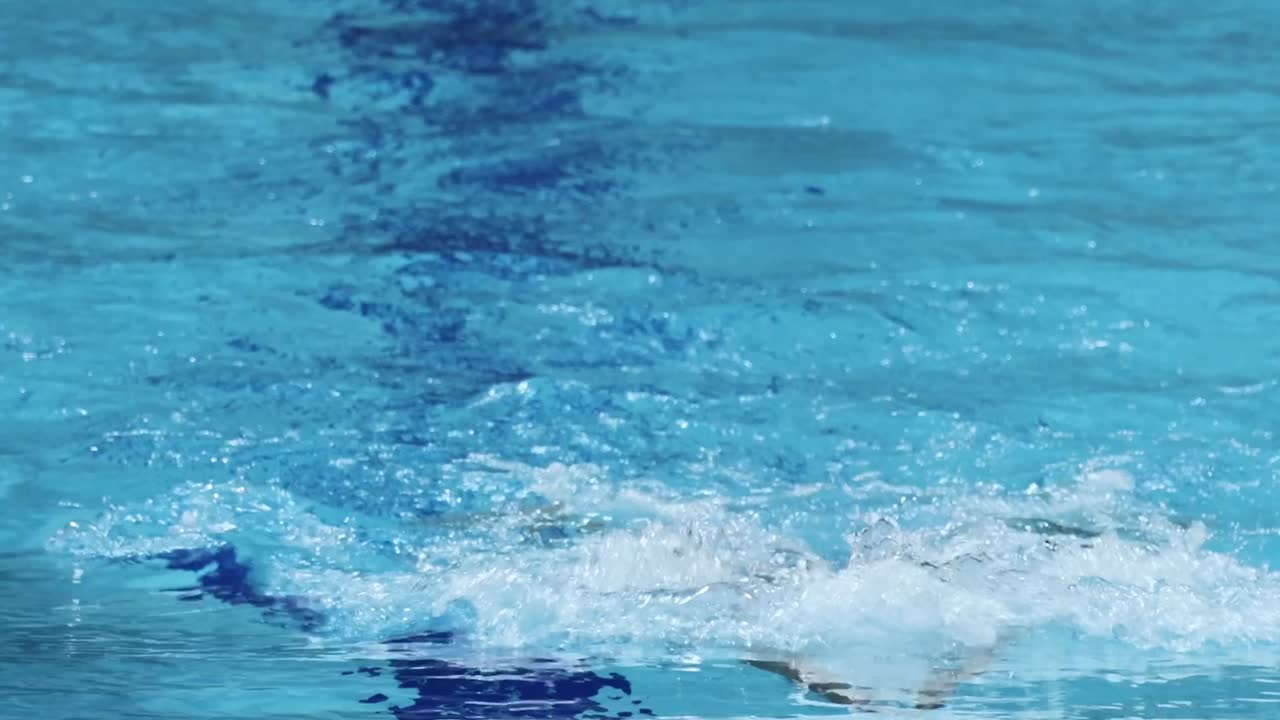Un atleta maschio nuota a farfalla verso la telecamera in una piscina di un blu brillante.