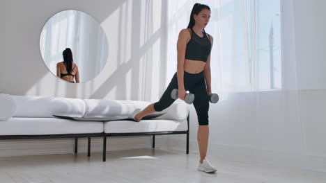 Brunette-in-black-sportswear-in-a-white-apartment-makes-a-split-squat-with-dumbbells-in-her-hands-leaning-on-the-sofa-with-her-foot.