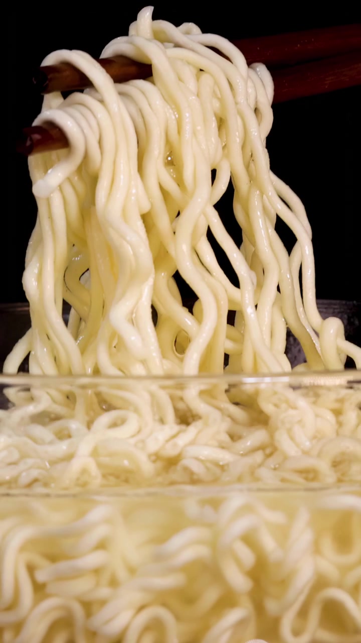 Bacchette di legno sollevano noodles istantanei cotti da una ciotola di vetro trasparente su uno sfondo nero, con illuminazione morbida e uniforme e movimento fluido della telecamera