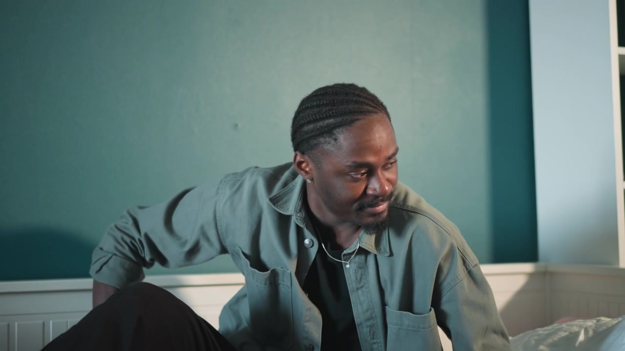 Black man sitting up on bed, preparing to reach for phone and call for help focused expression, steadying with hand on bedside, rising from blanket, home interior, determination to seek medical