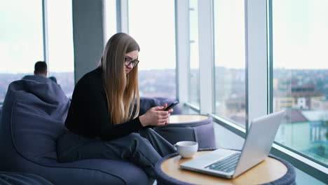 young woman writing sms on a phone. beautiful business woman sitting in a comfortable chair in front of a laptop and uses a mobile phone in light office center. profile view.
