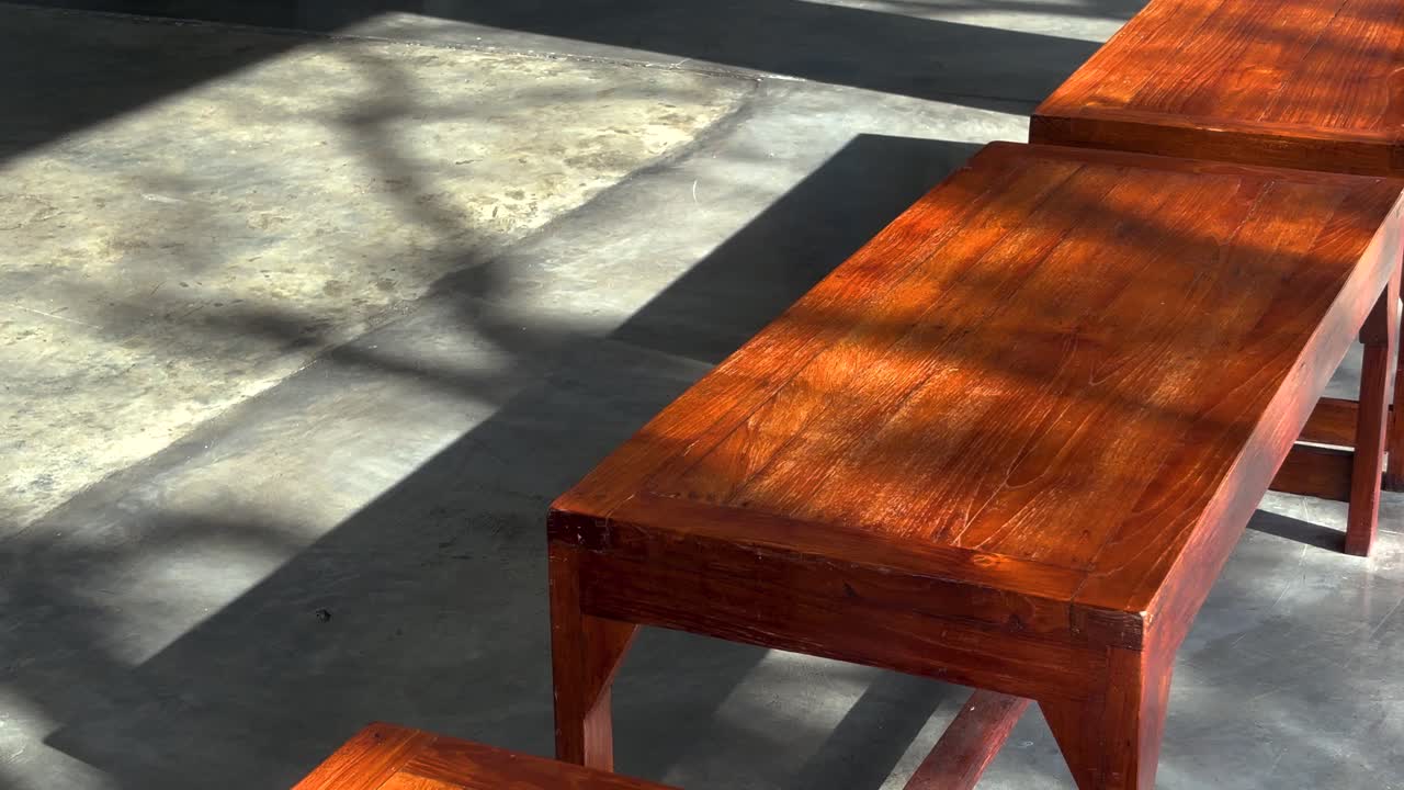 Un primo piano statico di una semplice panca di legno su un pavimento di cemento liscio. La calda luce del sole illumina la superficie rossastra dell'asse, mettendo in risalto le venature del legno.