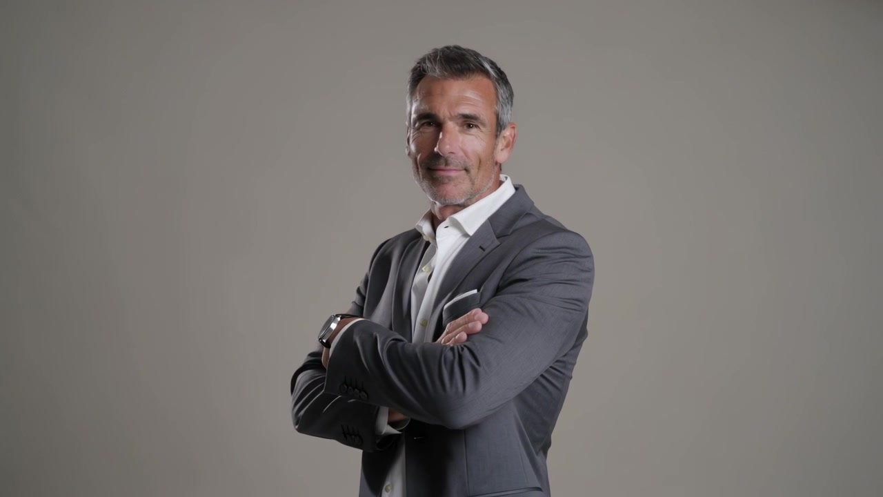 Un uomo in abito grigio cammina e si gira verso la telecamera dopo un segnale in studio, aggiustandosi l'orologio e incrociando le braccia.