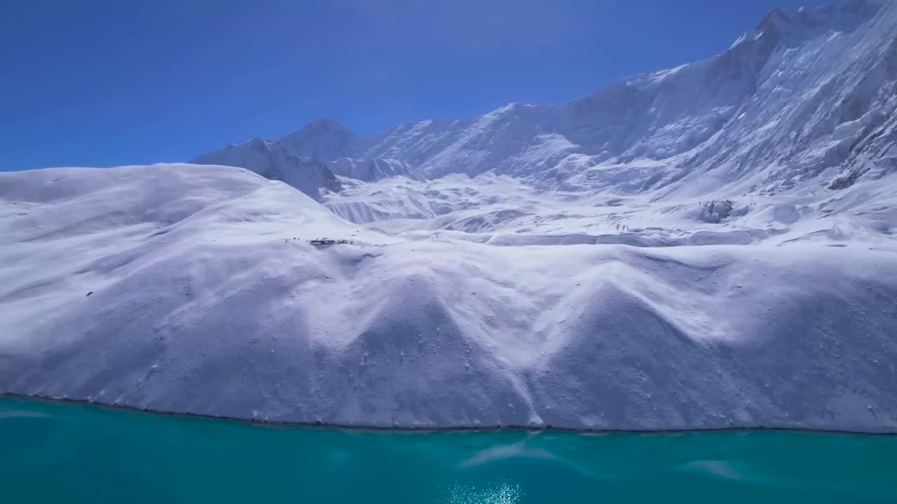 Aerial view of blue Tilicho Lake in Nepal. Drone slowly moves towards to lake shore and white snow-covered rugged landscape, snowy jagged mountain peaks in the horizon. Clear blue sky and sunny day