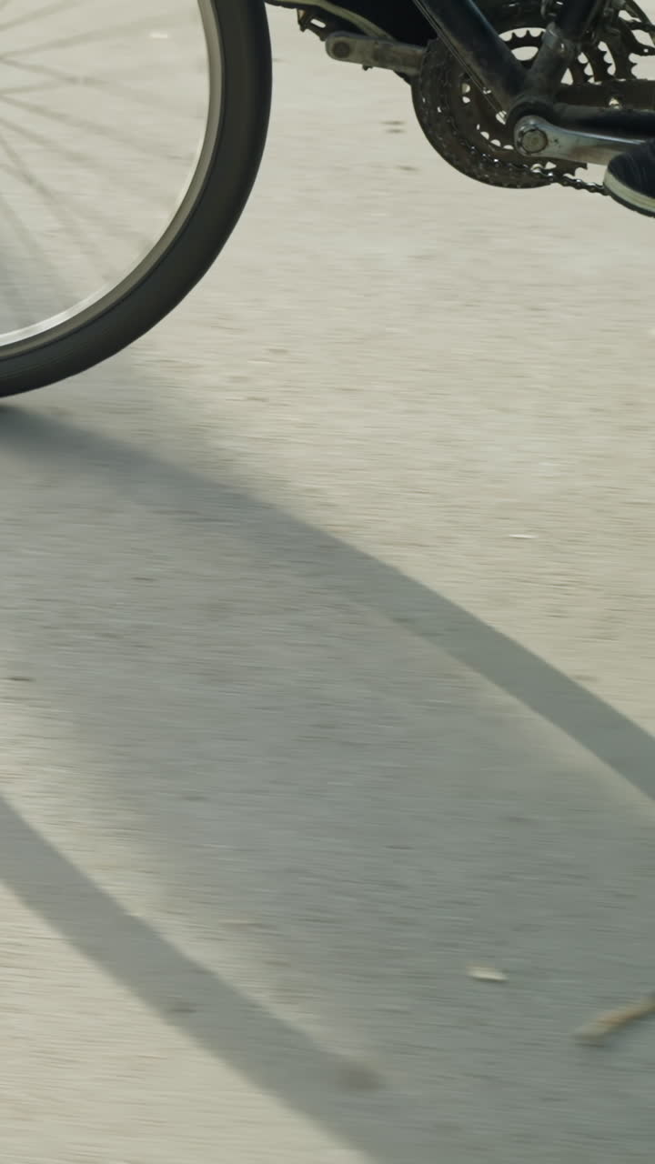 Leg view of someone wearing sneakers and black trousers, pedaling a bicycle slowly with a distinct shadow of the bike cast on the sunlit road