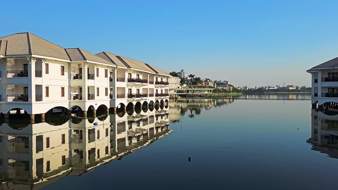 The InterContinental Hanoi Westlake Hotel features elegant white waterfront buildings with arched terraces reflecting perfectly in the tranquil lake waters under a clear blue Vietnamese sky